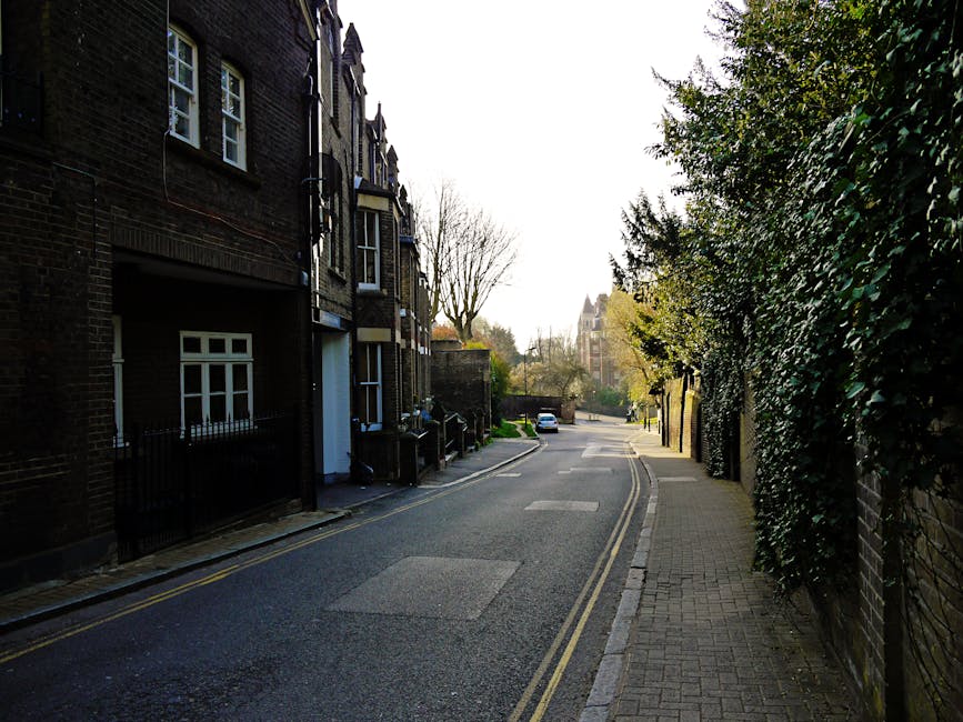 A narrow residential street in Bromley, with a pavement on the right side bordered by a tall hedge and a row of multi-storey brick houses on the left. The street has double yellow lines along the edge
